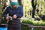 Man standing outdoors, wearing a face mask, working on a large metal garden fork or pitchfork with an angle grinder.