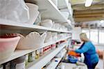 A woman working at a bench in a pottery studio. Shelves storing pots and bowls.