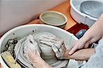 A person seated at a potter's wheel turntable, with hands on a wet clay lump, being taught to handle the clay.