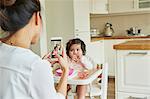 Over shoulder view of woman photographing baby daughter in high chair