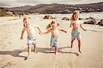 Three young sisters holding hands, running along beach, smiling