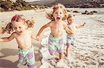 Three young sisters holding hands, running along beach, smiling