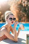 Young woman with dreadlocks, relaxing beside swimming pool, holding cold drink