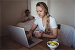 Woman using laptop while having coffee in bedroom