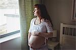 Thoughtful pregnant woman looking through window in bedroom