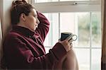 Thoughtful woman sitting at window sill and holding coffee cup