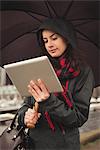 Woman looking at digital tablet while holding umbrella in city