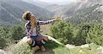 Young smiling woman with backpack sitting on rock taking selfie with beautiful views of mountains on background.