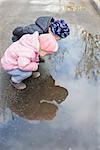 On an asphalt road, two children sit near a puddle and look at their reflection in it