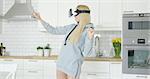 Young female wearing sweat shirt standing among kitchen and enjoying VR headset while gesticulating and laughing alone.