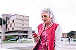 Mature woman in baseball jacket listening to headphones and looking at smartphone