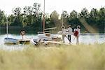 Friends walking towards sailing boat, carrying equipment, long grass in foreground