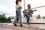 Three friends relaxing on pier, sitting on sailing boat, man playing guitar