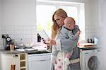 Mother standing in kitchen, holding baby boy, looking at digital tablet