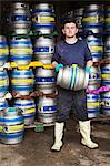 Man working in a brewery, standing next to a stack of metal beer kegs, holding beer keg.