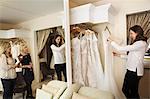 Three women, a client and two retail advisors in a wedding dress shop, looking through the choice of gowns. Large mirror and rows of bridal gowns.