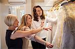 Three women in a wedding dress boutique looking at a rail of white bridal gowns, with lace and decorative fabrics. A bride to be and two sales assistants.