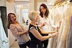 Three women in a wedding dress boutique looking at a rail of white bridal gowns, with lace and decorative fabrics. A bride to be and two sales assistants laughing.