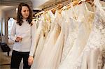 Rows of wedding dresses on display in a specialist wedding dress shop. A woman holding a glass of champagne looking at a rack of couture gowns with lace and net.