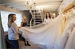 Three women in a wedding dress shop, one holding out the full skirt of a bridal gown.