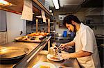 Chef standing in a restaurant kitchen at a counter, holding sauce pan, plating food.