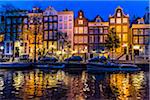 Traditional buildings illuminated in the evening with parked cars and a boat touring along the Herengracht Canal in Amsterdam, Holland