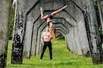 Young woman balancing on top of man hands, practicing yoga below concrete bridge