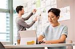 Female digital designer typing on laptop and colleague arranging mood board in office