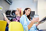 Two women reading newspapers back to back in laundrette