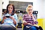 Two women sitting in laundrette reading magazine and looking at smartphone