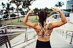Young woman outdoors, standing at top of steps, flexing muscles, rear view, South Point Park, Miami Beach, Florida, USA
