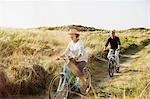 Playful mature couple riding bicycles on sunny beach grass path