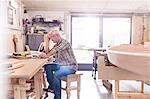 Male carpenter working at laptop on workbench near wood boat in workshop