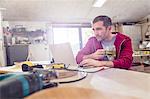 Male carpenter drinking tea and working at laptop on workbench in workshop