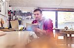 Male carpenter using digital tablet, drinking tea at workbench in workshop