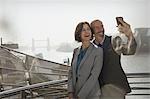 Enthusiastic, smiling business couple taking selfie with camera phone on sunny urban bridge, London, UK