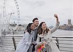 Smiling, happy friends taking selfie with selfie stick on bridge near Millennium Wheel, London, UK