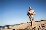 Bare chested young male runner running along beach against blue sky