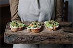 Woman holding chopping board, with ricotta, avocado and walnut bruschetta on top, mid section