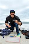 Young male sea fisher looking at hook while crouching on beach