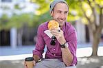 Young man sitting outdoors, holding takeaway coffee cup and croissant