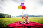 Girl sitting on red blanket in rural field looking up at bunch of colourful balloons