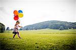 Girl walking in rural field with bunch of colourful balloons