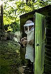 A beekeeper walking out of a shed wearing a veil and holding a smoker to calm honeybees.