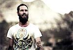 Portrait of bearded man wearing printed T-Shirt, tattoos on arms and chest, mountains in the background.