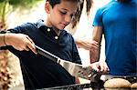 A boy holding tongs and turning food on a barbecue.