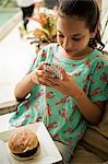 A seated girl with a burger on a plate in her lap, looking at a mobile phone.