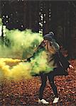 A young woman twirling with a yellow smoke flare in a forest.