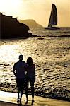 Rear view of couple standing on a sandy beach by the ocean, cliffs and a sailing boat in the distance at sunset.