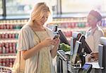 Smiling young woman texting with cell phone at grocery store market checkout